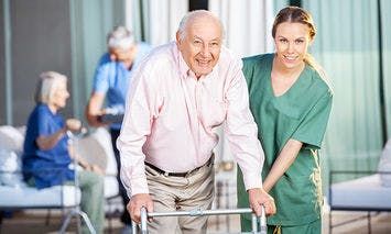 An elderly man using a walker is assisted by a caregiver in green scrubs, smiling together in an outdoor setting with other elderly individuals and caregivers in the background.